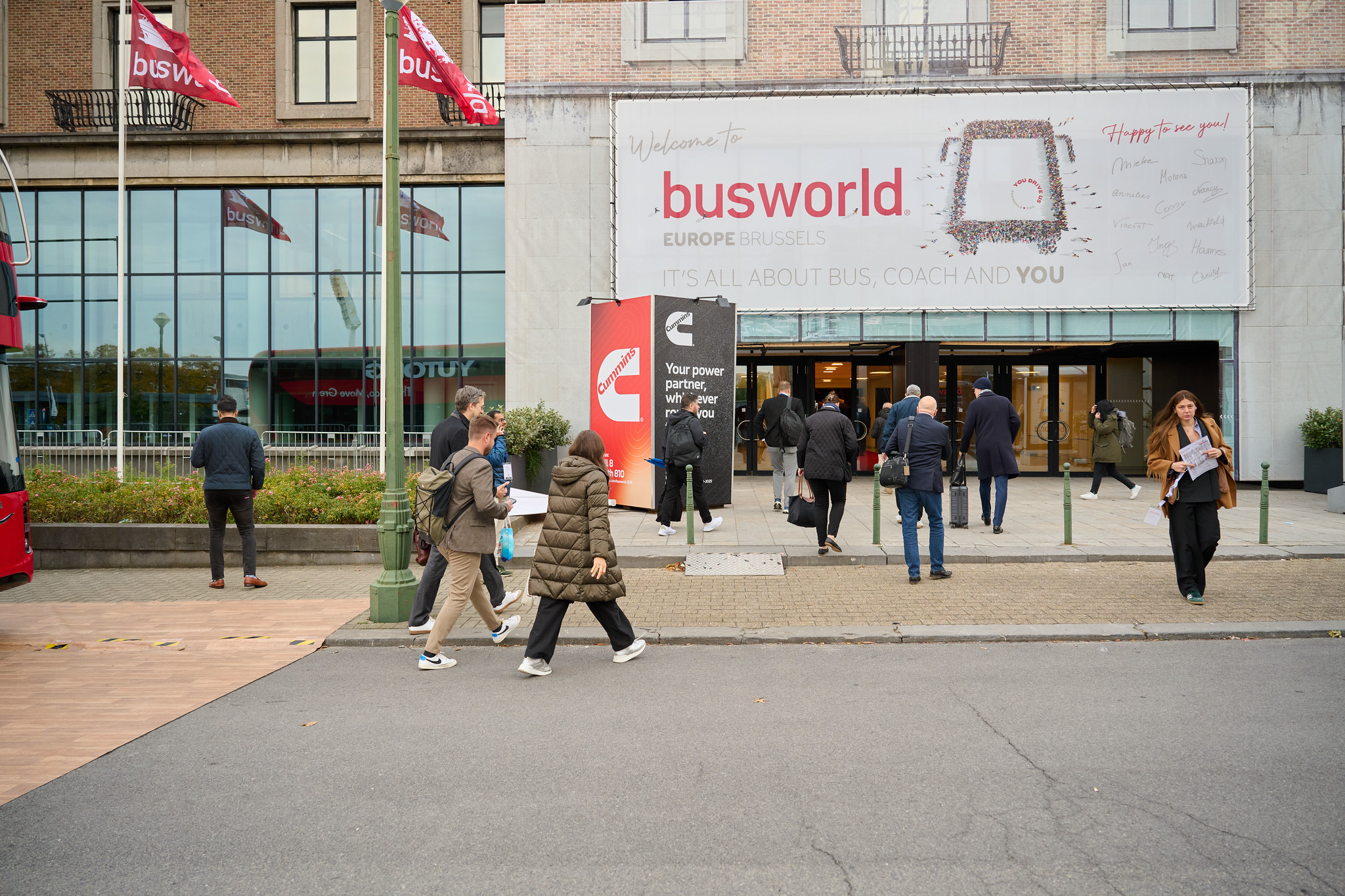 Visitors arriving at Busworld Europe 2025 in Brussels, Belgium, with Busworld flags and a welcome banner at the entrance to the international bus and coach exhibition.